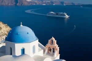 A cruise ship plies the waters of Greece with a white-washed blue-domed building in the foreground, high above